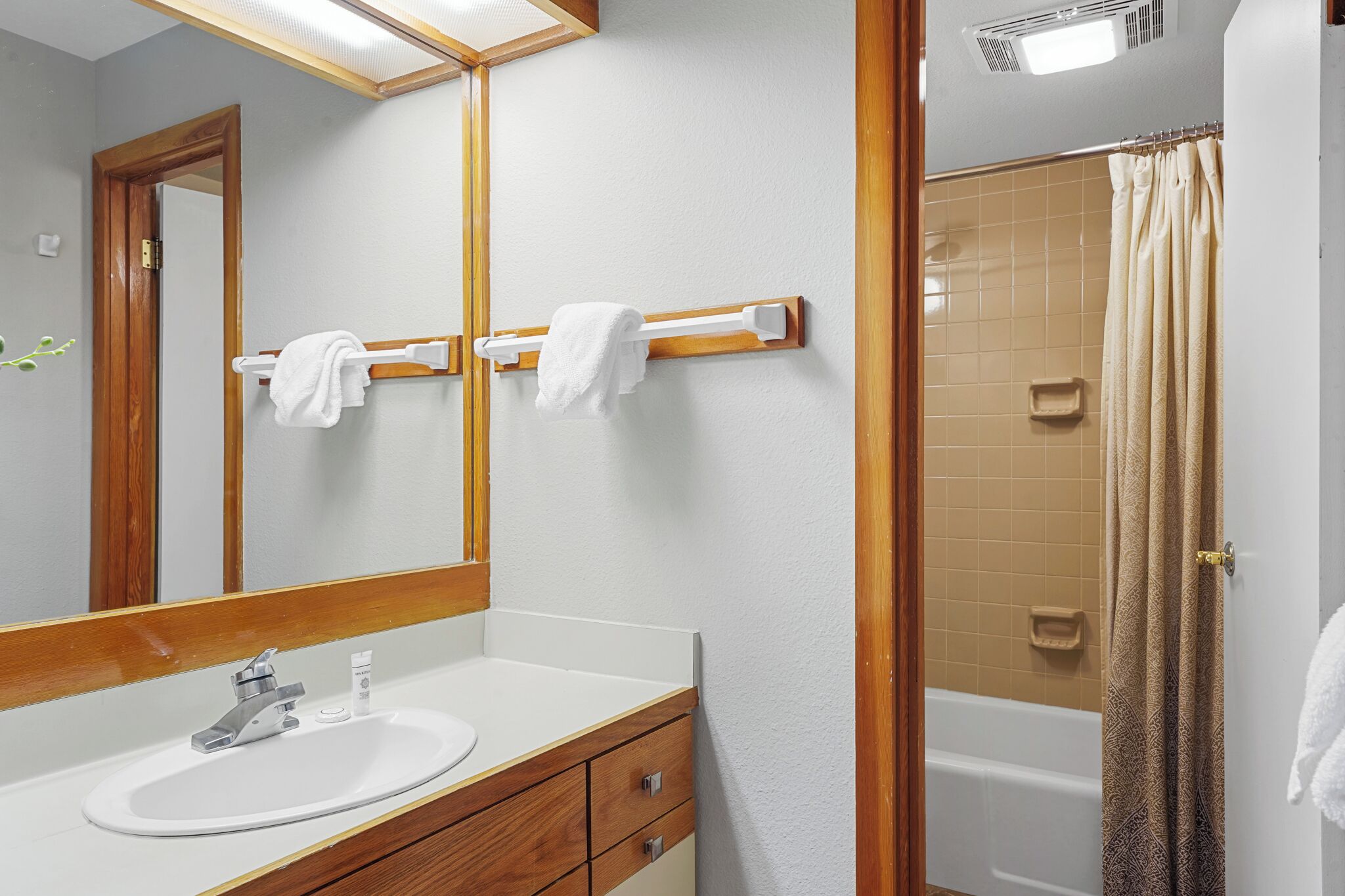 Une salle de bain avec un meuble-lavabo en bois, un évier, un miroir, deux porte-serviettes et une douche avec un rideau de couleur beige.