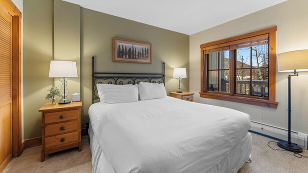 A neatly arranged bedroom with a large bed, two bedside tables with lamps, a framed picture on the wall, and a window with a view. - Expedition Station #8582 (Keystone)