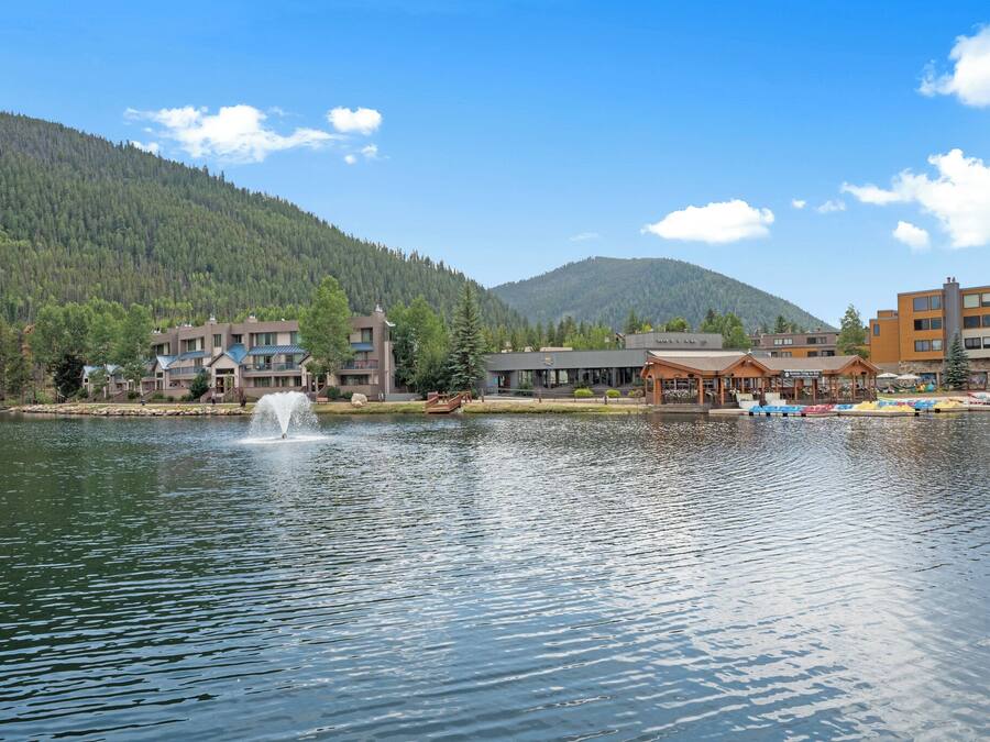 A lakeside view with buildings, a fountain, and a forested mountain in the background.