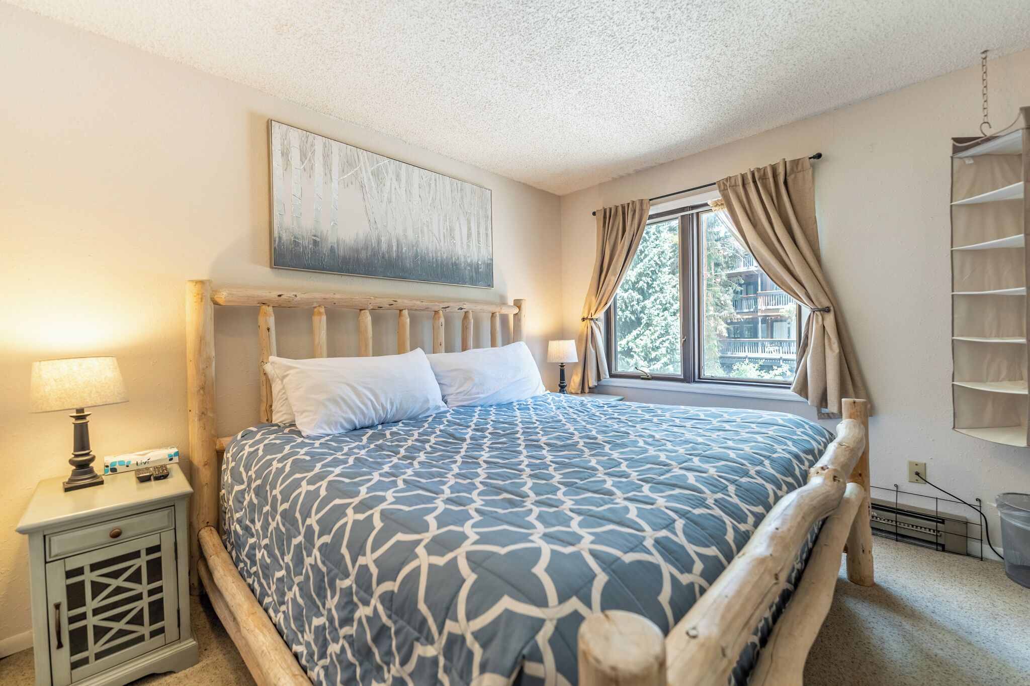 A bedroom with a wooden bed frame, a patterned bedspread, a nightstand with a lamp, and a window with curtains.