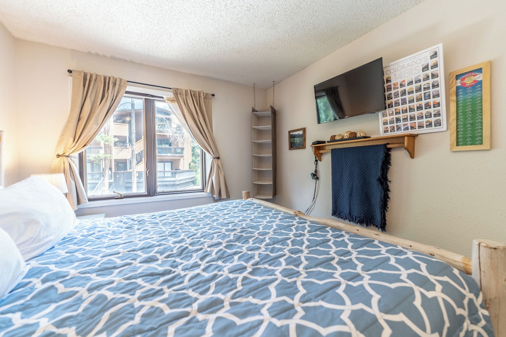 A bedroom with a bed, a window with curtains, a TV, a shelf with items, and a wall with a calendar and picture.