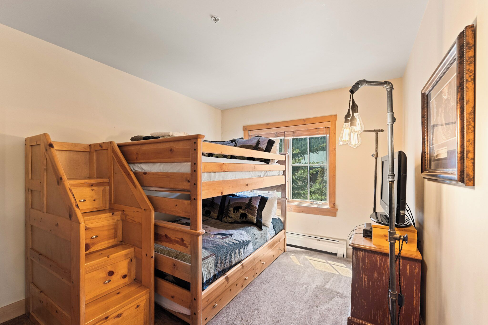 A room with a bunk bed, wooden staircase, a desk with a lamp, and a window with blinds.