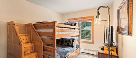 A room with a bunk bed, wooden staircase, a desk with a lamp, and a window with blinds.