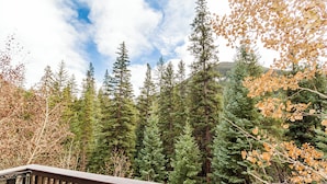 A wooden deck with a metal table, surrounded by dense evergreen trees and a backdrop of a mountain range.