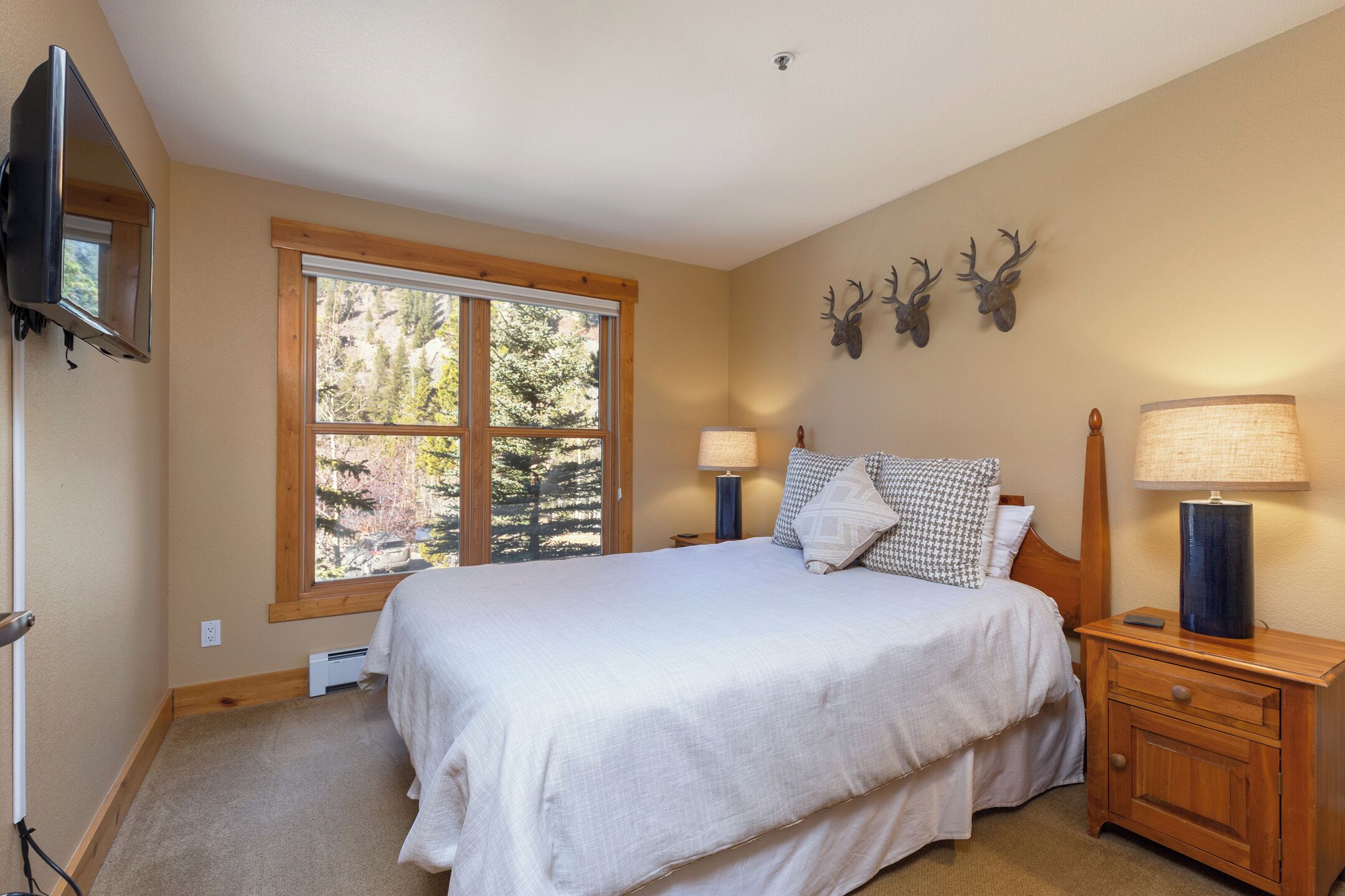 Une chambre avec une grande fenêtre, un lit recouvert de draps blancs, deux lampes de chevet, une table de nuit en bois et une paire de bois de cerf fixée au mur.