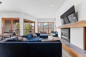 A modern living room with a large sectional sofa, a fireplace, and large windows offering a view of the outdoors. - Qs1645-2 (Keystone)