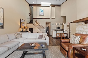 A modern living room with a sectional sofa, a leather armchair, and a coffee table. A staircase leads to a kitchen area with bar stools.