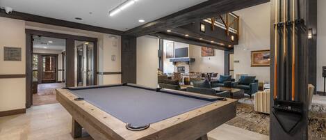 A pool table in a modern living area with a fireplace, comfortable seating, and a staircase leading to an upper level.