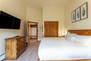 Un dormitorio con cama, cómoda de madera, televisor, armario y una obra de arte con hojas enmarco.