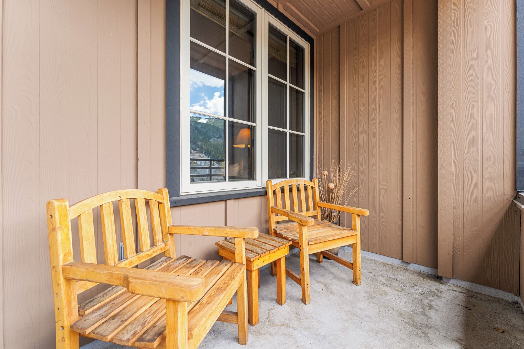 A wooden porch with two wooden chairs and a table, a large window, and a view of trees and a cloudy sky.