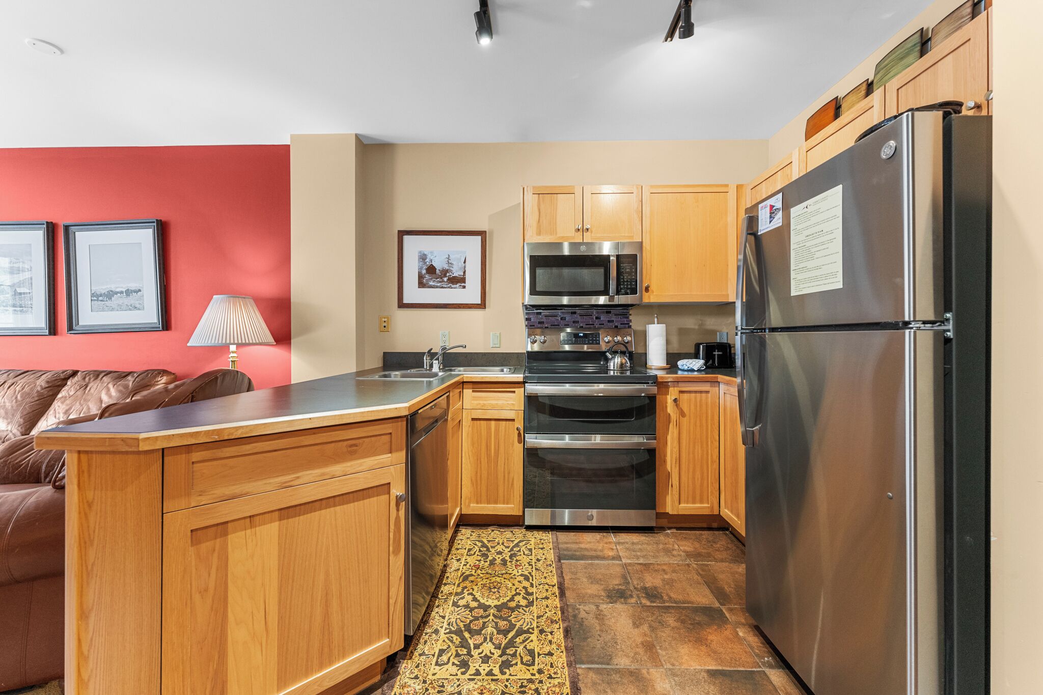 A compact kitchen with wooden cabinets, a stainless steel refrigerator, a microwave, and an oven. There is a rug on the floor and a framed picture on the wall.