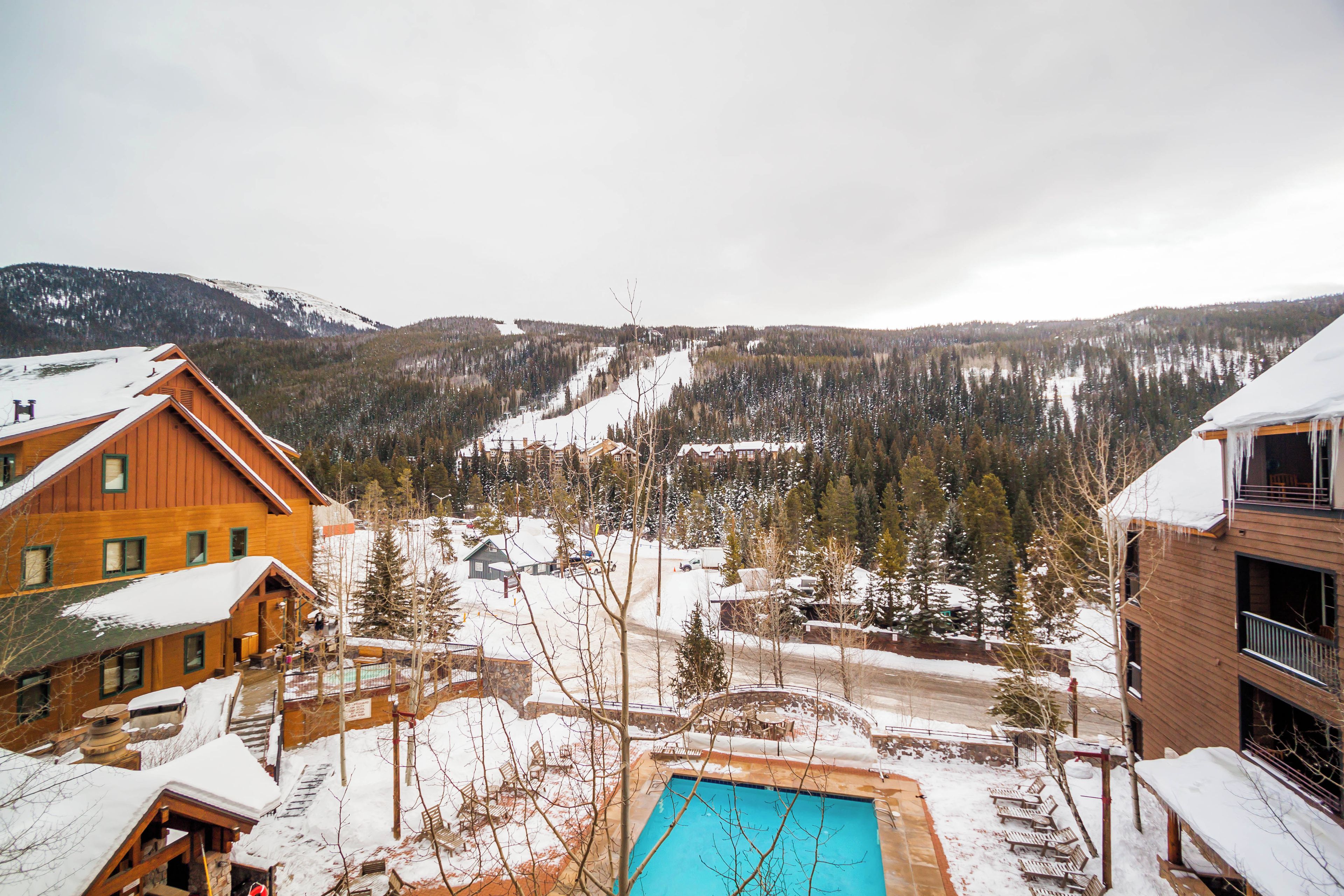 Una cabaña de madera con piscina, rodeada de árboles nevados y montañas.