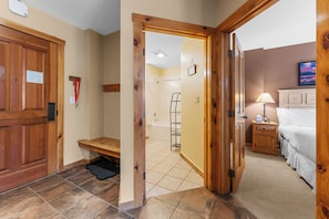 Habitación de hotel con puerta de madera, un banco, ducha, cama, mesita de noche y lámpara.