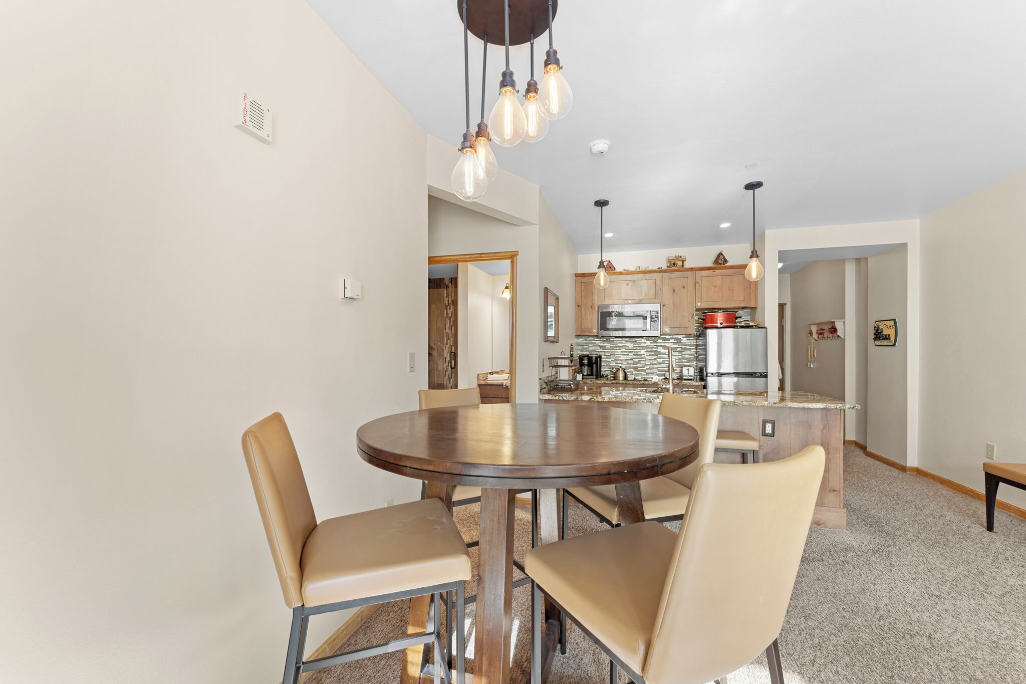 A dining area with a round table and chairs, a kitchen in the background, and pendant lighting.