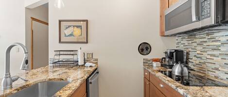 A kitchen with granite countertops, wooden cabinets, a stainless steel sink, a microwave, and a coffee maker.