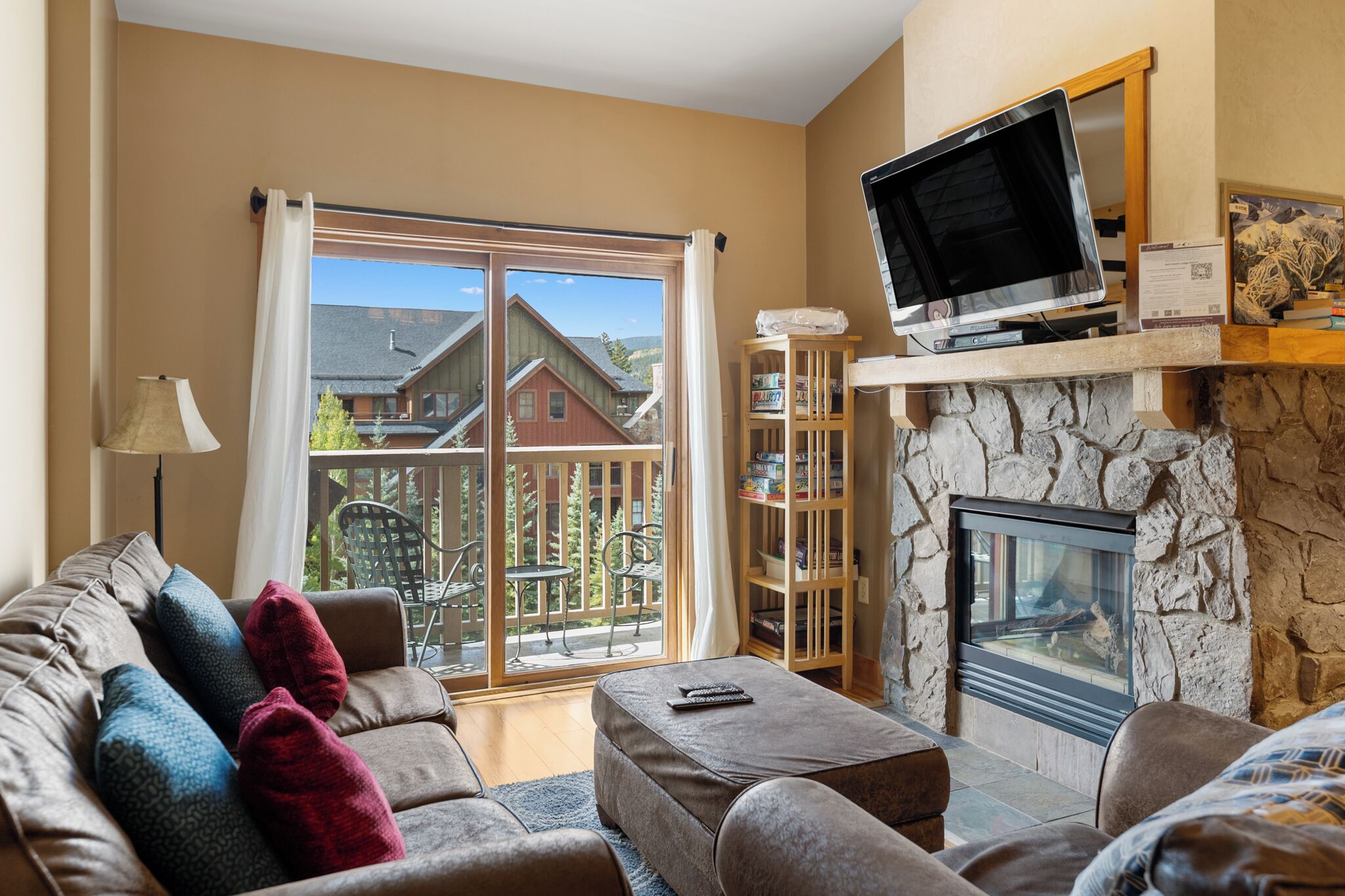 A cozy living room with a stone fireplace, a large window with curtains, a sofa with cushions, and a television on a shelf.