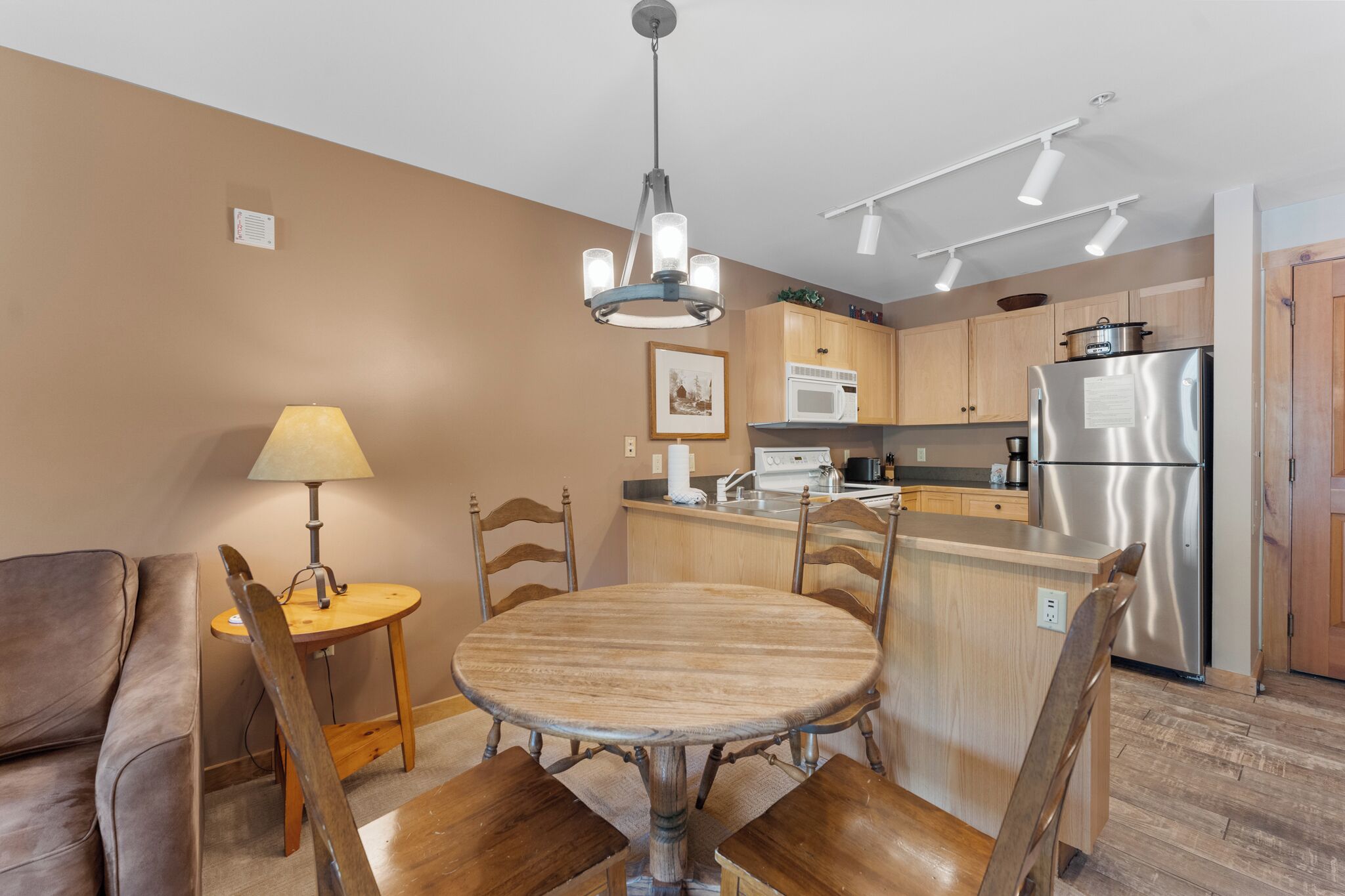 A compact kitchen with a central island, wooden chairs, a dining table, a lamp, and modern appliances.