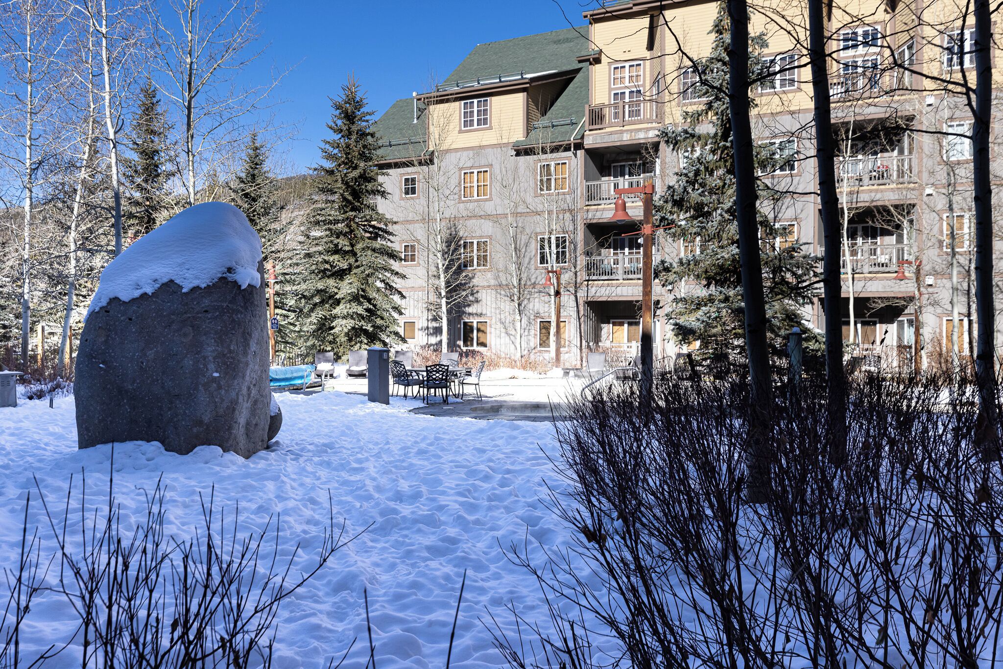 A snow-covered outdoor area with a large rock, trees, and a multi-story building with balconies.