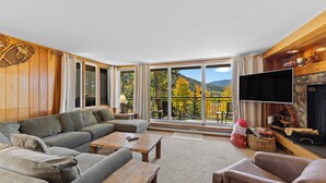 Ein geräumiges Wohnzimmer mit einer großen Couch, einem Couchtisch und einem Kamin. Ein großes Fenster bietet Blick auf Bäume und Berge.