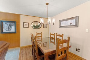 Une salle à manger avec une table à plateau de verre, des chaises en bois, un lustre, des tableaux encadrés et un miroir au mur.