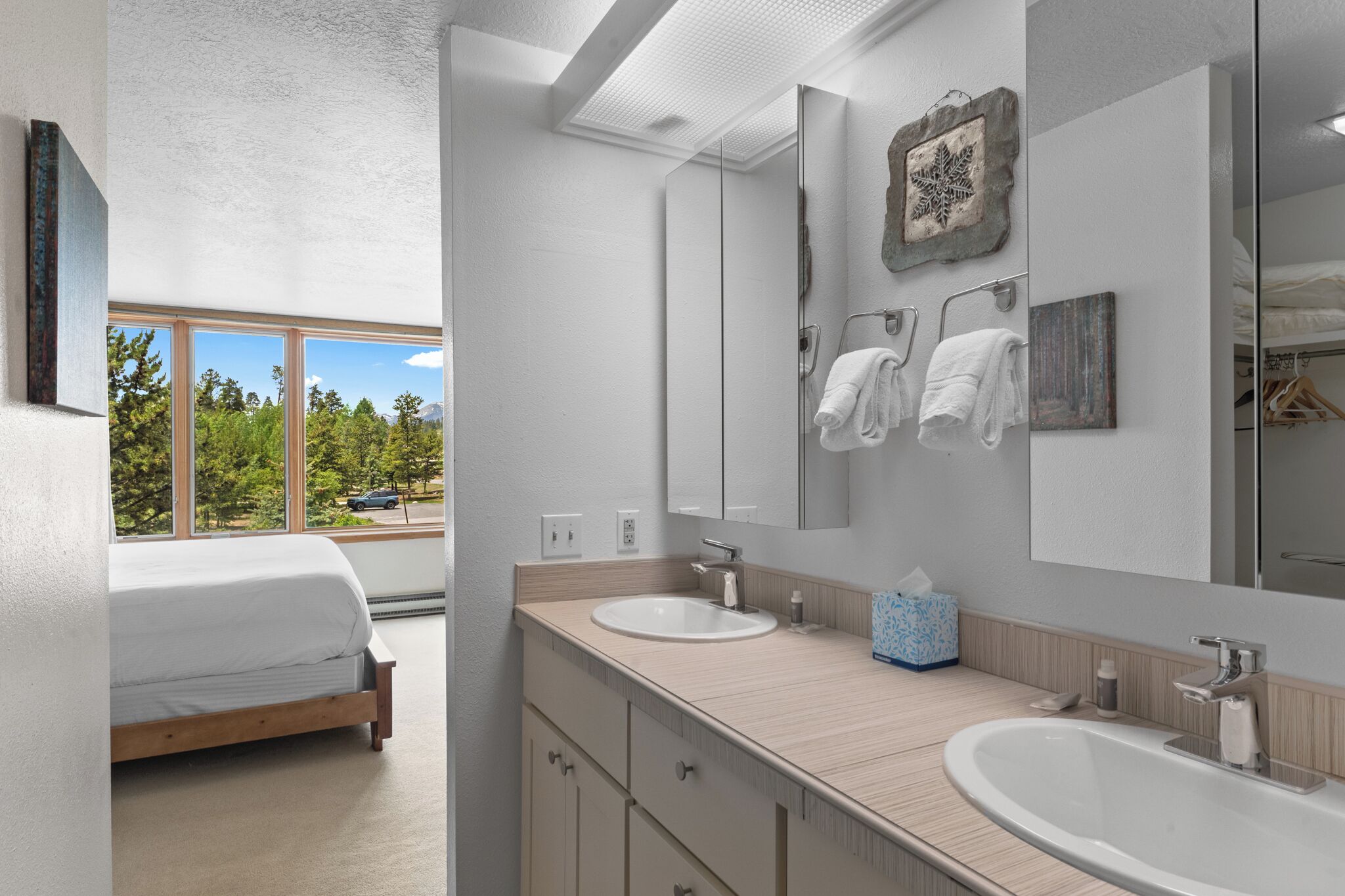 A modern bathroom with two sinks, a large mirror, and a window with a view of trees and a car.