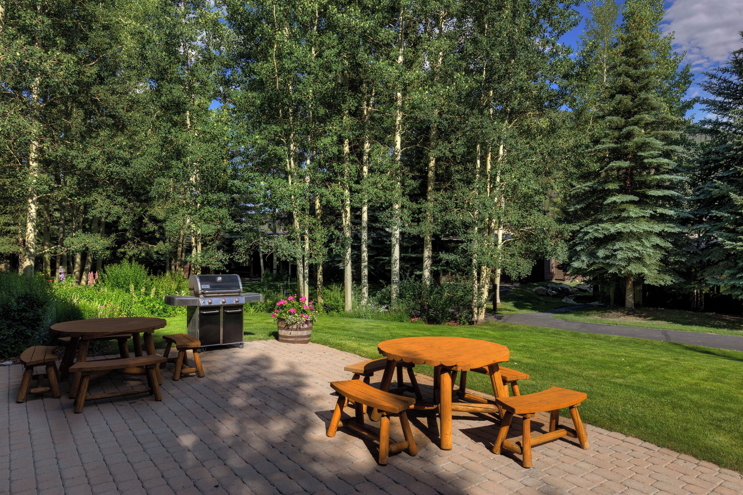 A wooden picnic table set on a brick patio surrounded by lush greenery and tall trees.