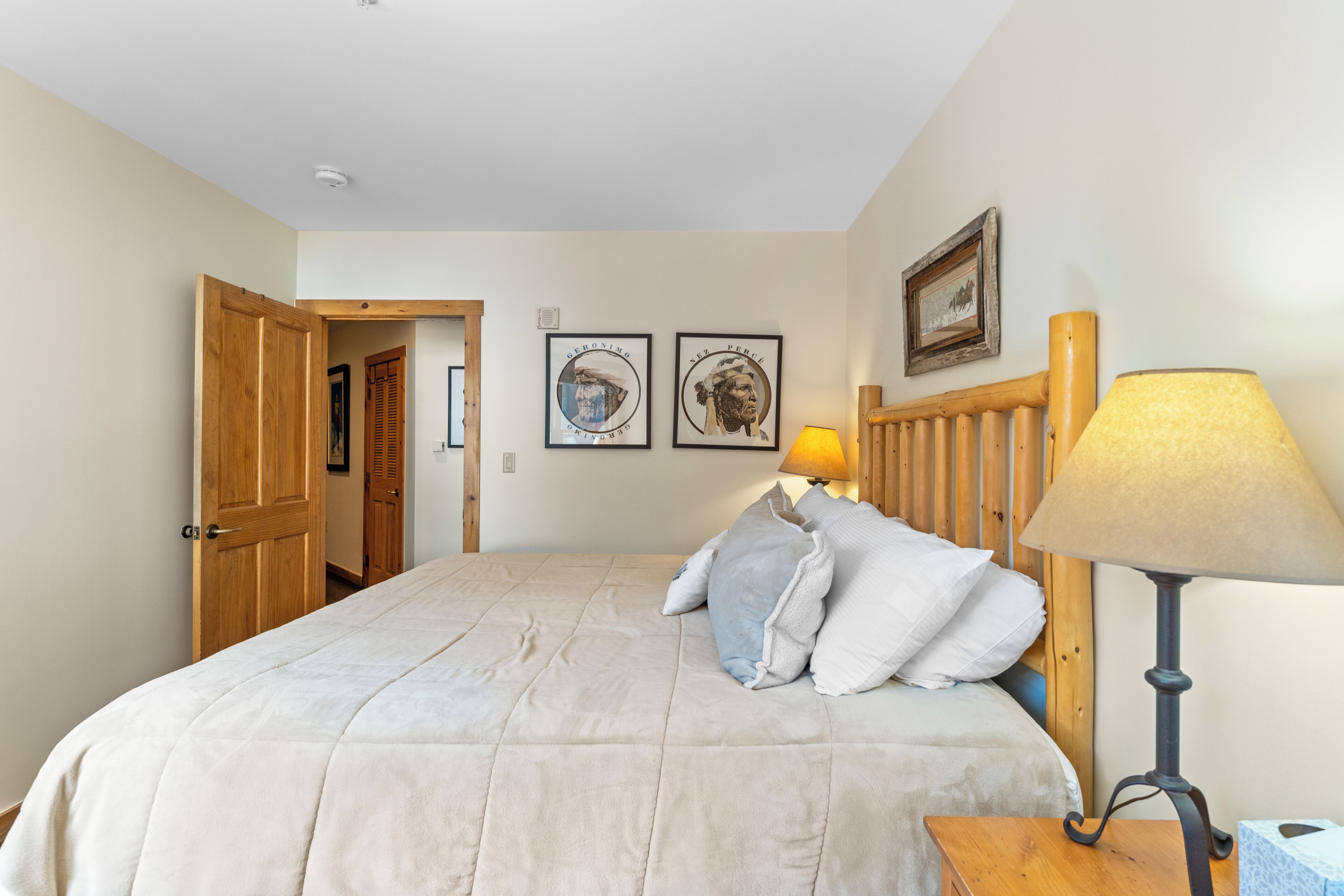 A bedroom with a bed, pillows, a wooden headboard, a bedside lamp, and two framed pictures on the wall.