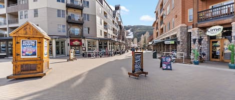 A paved pedestrian area with multi-story buildings, a wooden kiosk, and a signboard.