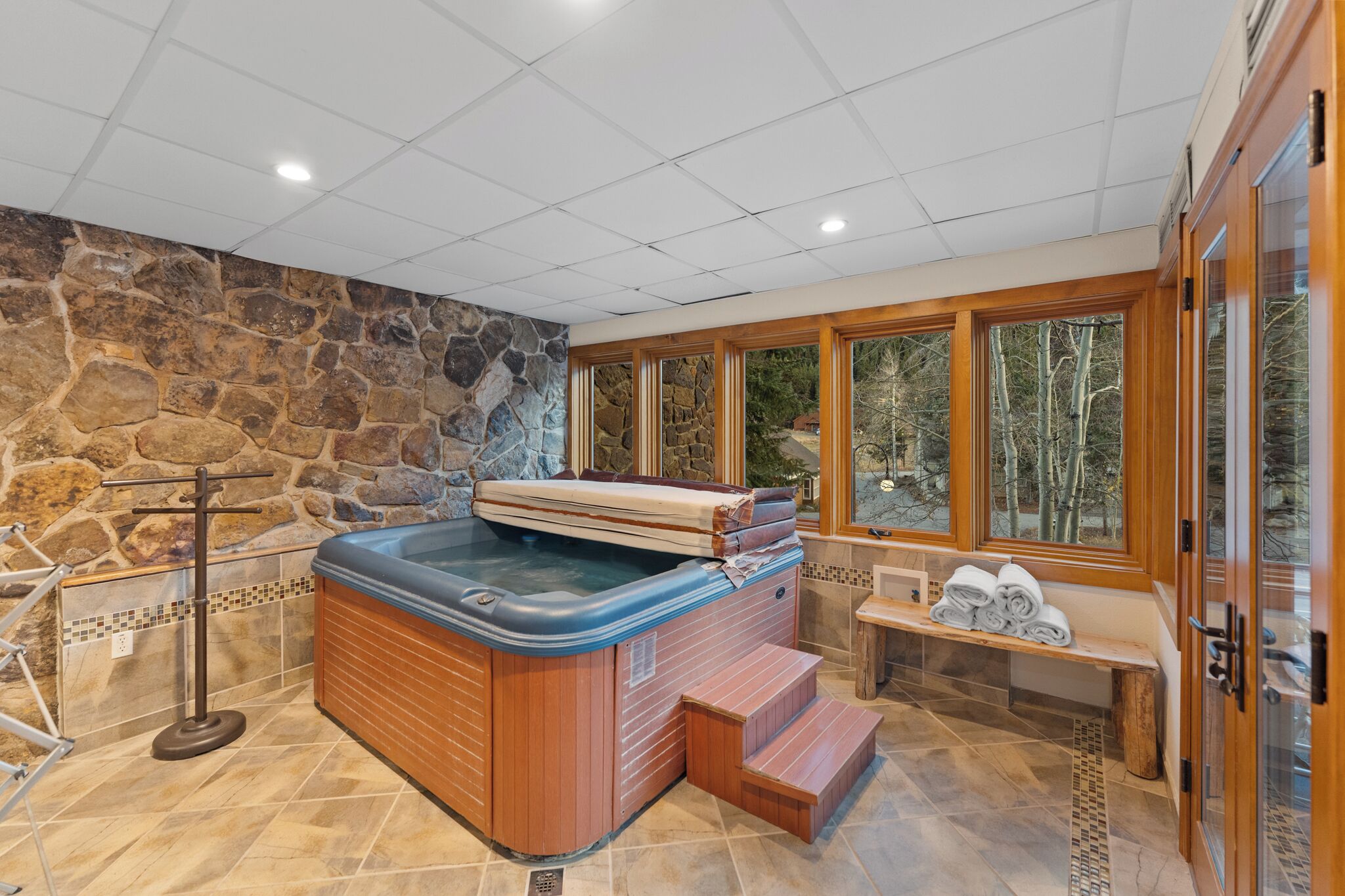 A hot tub with wooden steps, a stone wall, and a bench with towels.