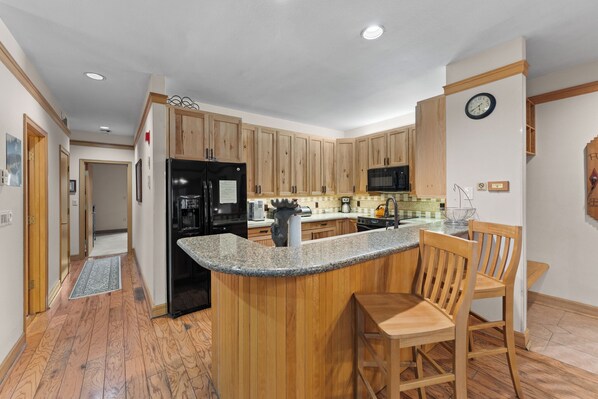 A kitchen with wooden cabinets, a granite countertop, a black refrigerator, a microwave, and a dining area with a wooden table and chairs.