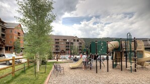 A playground with a slide and climbing structures, surrounded by a gravel area and a wooden fence. - Dakota Lodge #8517 (Keystone)