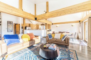 A spacious living area with high ceilings and exposed wooden beams, featuring two sofas with colorful cushions, a coffee table, and a rug.