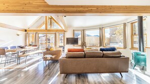 A spacious living area with a large sofa, wooden beams, and large windows offering views of the outside.
