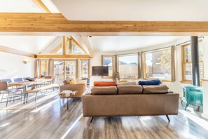A spacious living room with a large sofa, a television, and a fireplace. There are multiple windows providing natural light and views of the outside.