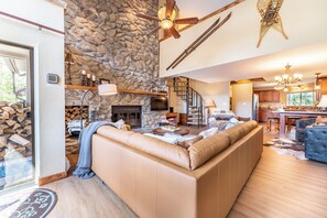 Amplia sala de estar con una pared de piedra, una chimenea, un sofá seccional beige grande y una cocina al fondo.