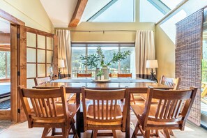 A dining area with a large wooden table and chairs, a vase with greenery, and a view of the outdoors through large windows.