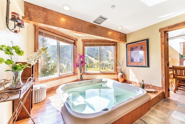 A room with a hot tub, large windows, wooden floors, and a view of mountains.