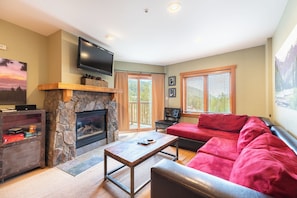 A living room with a stone fireplace, a red sofa, a television mounted above the fireplace, and a large window with blinds.