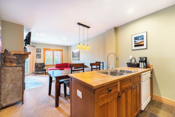 A compact kitchen with wooden cabinets, a sink, and a dishwasher. There is a dining area with a table and chairs, and a red sofa.