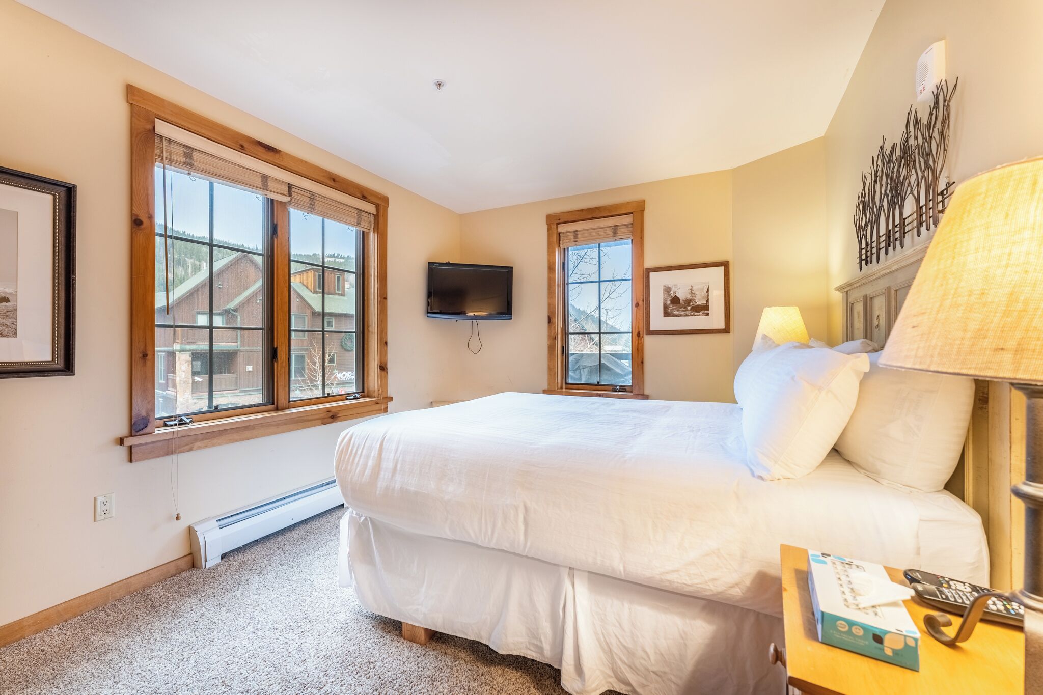 A bedroom with a large bed, two windows, a mounted TV, and a nightstand with a lamp.