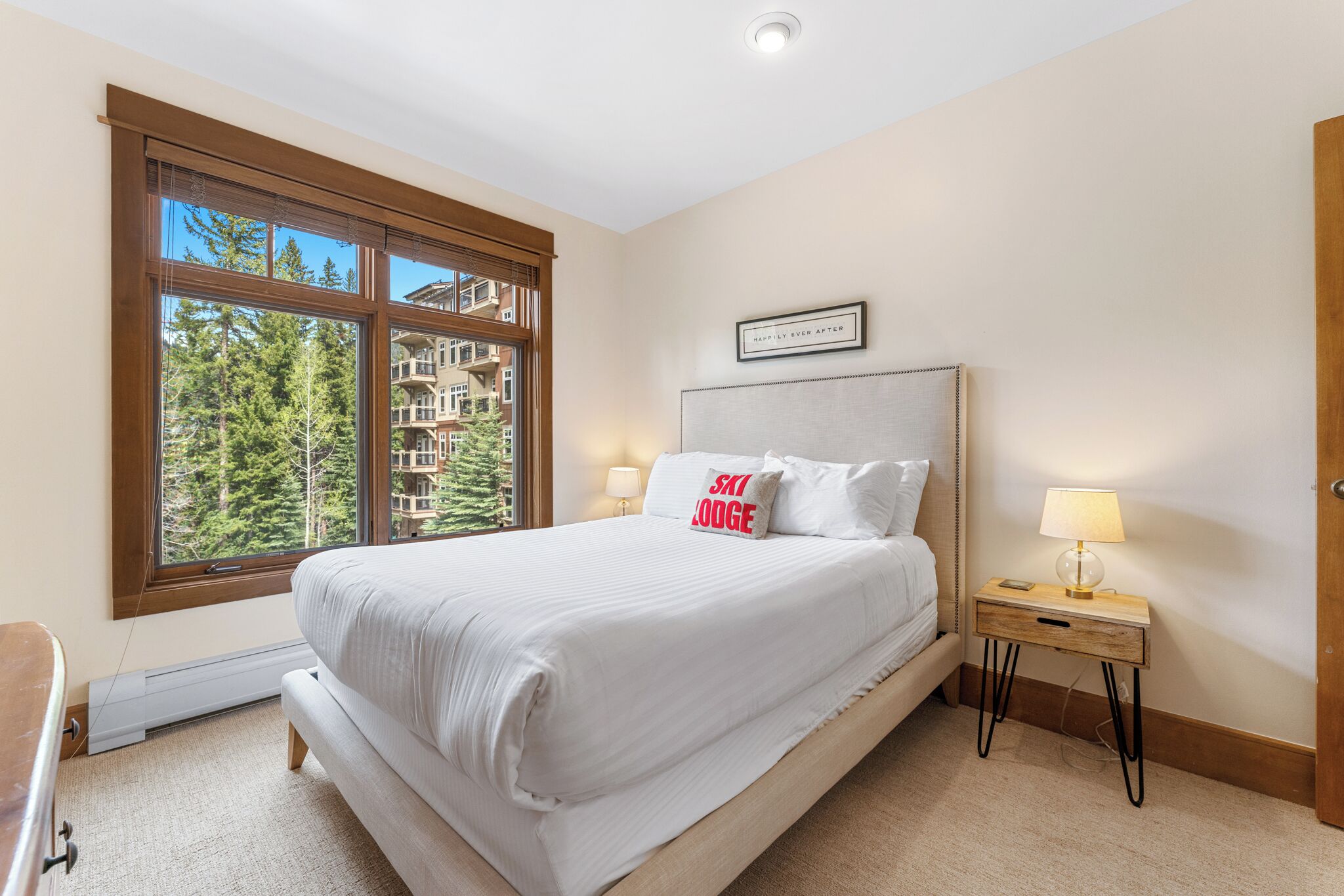 Une chambre avec une grande fenêtre, un lit recouvert d’une literie blanche, une table de chevet avec une lampe et une citation encadrée accrochée au mur.