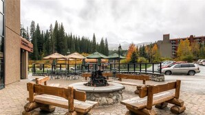 An outdoor seating area with wooden benches and tables, a central fire pit, and umbrellas.