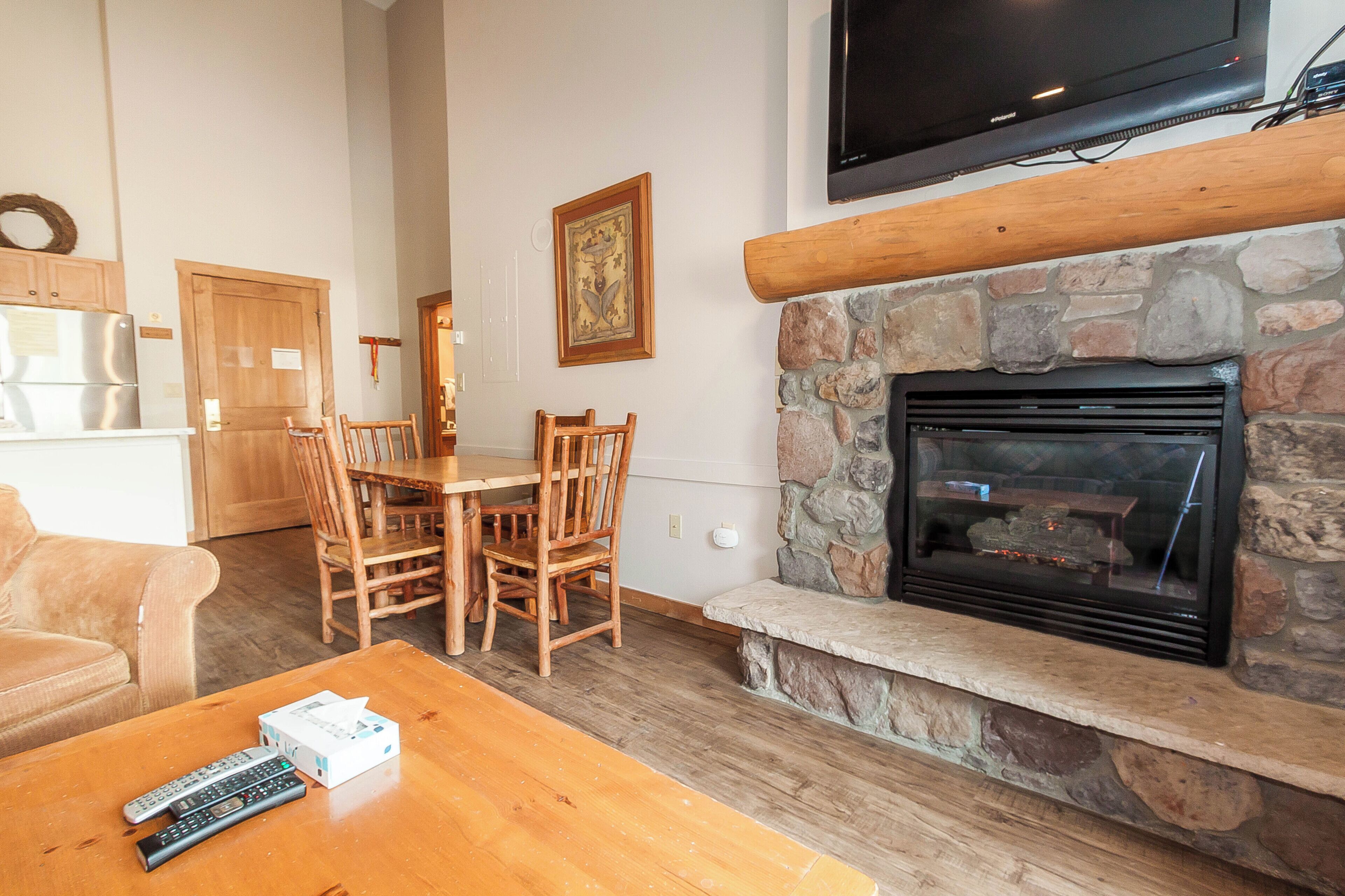 A living room with a stone fireplace, wooden furniture, a television mounted above the fireplace, and a kitchen area in the background.