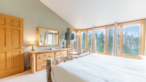 Ein Schlafzimmer mit einem großen Bett, einer Holzkommode, einem Fernseher und Blick auf Bäume und Häuser durch große Fenster.