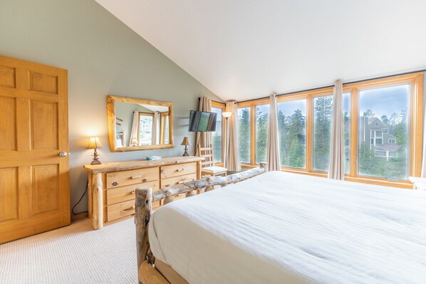 Une chambre avec un grand lit, une commode en bois, une télévision et une vue sur les arbres et les maisons, grâce à de grandes fenêtres.