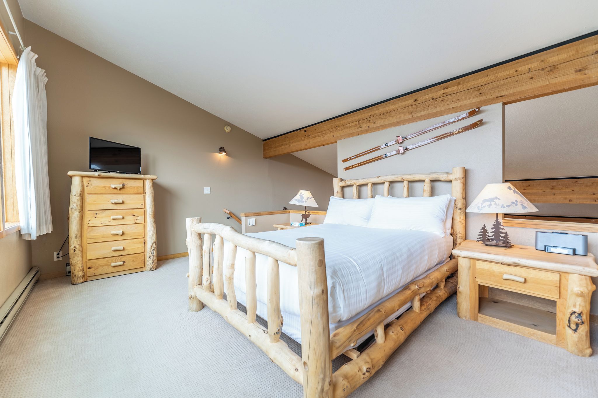 Een houten bed met hoofdeinde, een nachtkastje met lamp, een houten dressoir en een televisie op een dressoir.