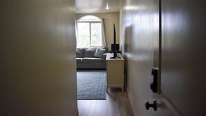 En smal gang fører til et rum med et stort vindue, en sofa og et skrivebord med en computer.
