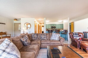 A living room with a brown leather sofa, a wooden coffee table, and a leather armchair. A kitchen is visible in the background with green cabinets and a microwave.