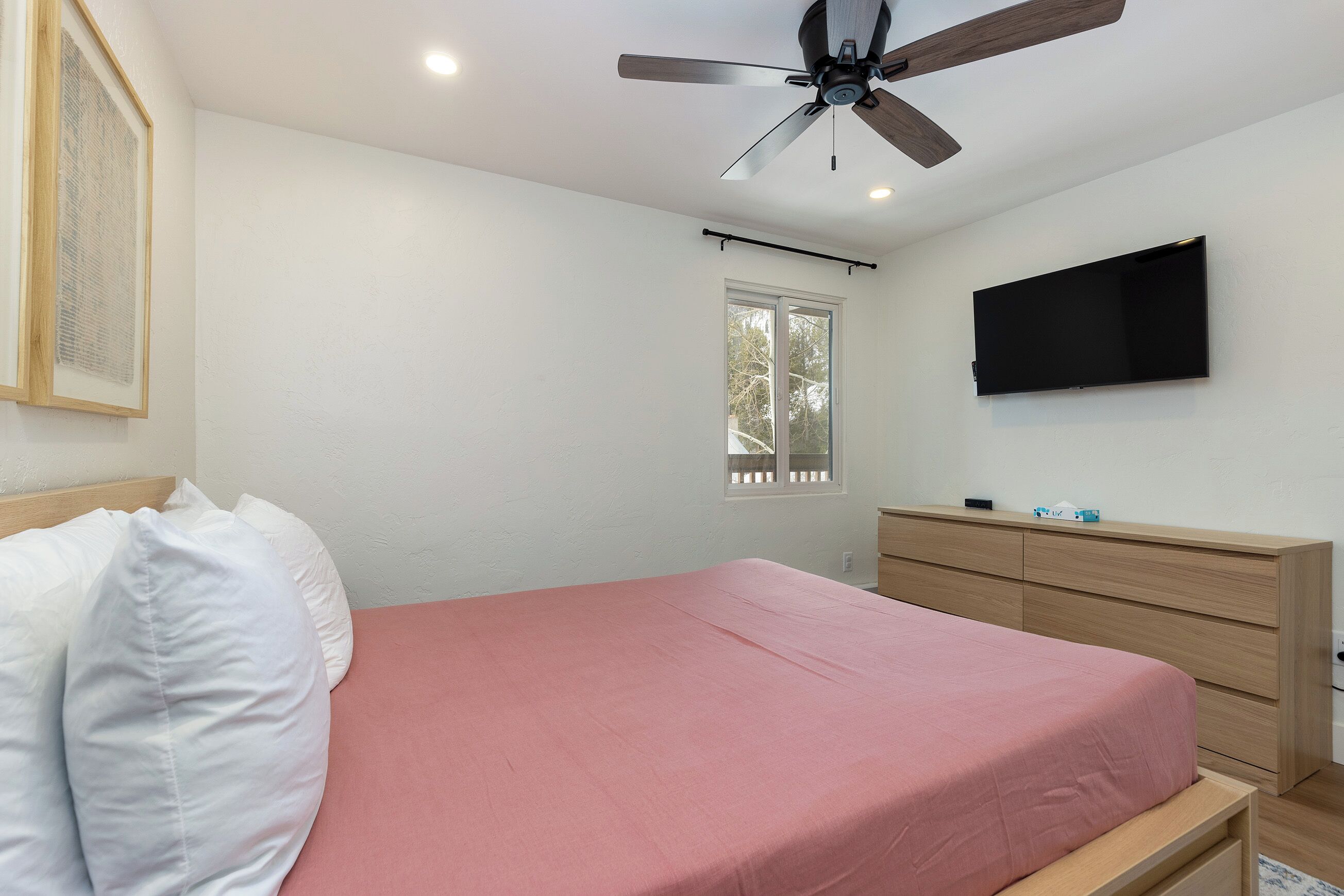 A bedroom with a bed, a ceiling fan, a mounted TV, a window, and a wooden dresser.