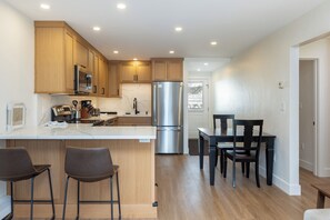 A modern kitchen with wooden cabinets, stainless steel appliances, a marble backsplash, and a dining area with a black table and chairs.